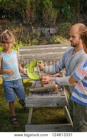 Father shows his sons how to make a sailing boat from pieces of wood and a piece card. They're outdoors