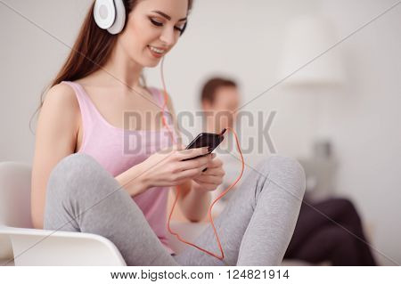Involved in hobby. Cheerful smiling attractive girl  holding cellphone and listening to music while her boyfriend sitting on the background