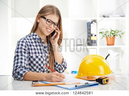 Young female architect consultant talking on phone in the office. Woman designer working on blueprint of industrial project.