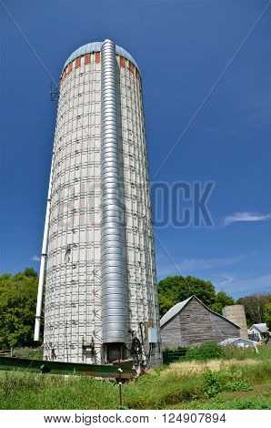 An unused large old silo rises into the sky overwhelming the weathered distant building in the background