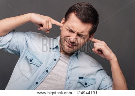 Too loud! Young handsome man covering ears by hands and keeping eyes closed while standing against grey background