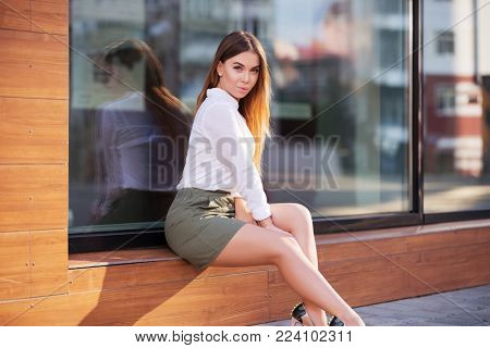 Young fashion woman sitting on the mall window in city street. Stylish female model wearing white blouse and short skirt outdoors