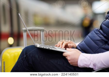 Freelancer working with a laptop in a train station while is waiting for transport. Middle age man on on railway station platform.