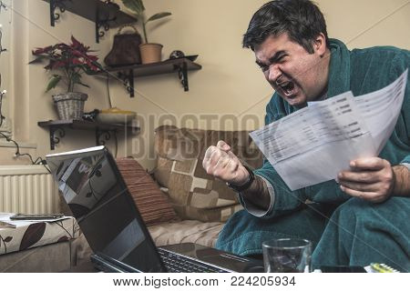 Enraged mature man yelling at his bills and financial reports, dressed in bathrobe, sitting in his living room