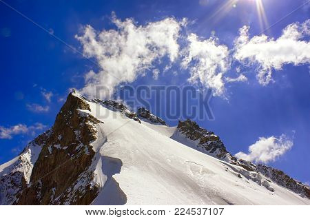 Closeup icy and snowy mountain peak with cliff ridge under blue sky with cloud and sunbeams. Sunny day.