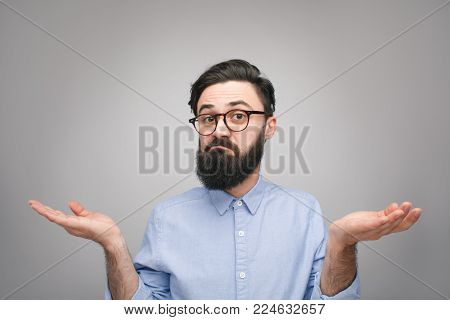 Young bearded man in bewilderment shrugging his shoulders and looking at camera with confusion.