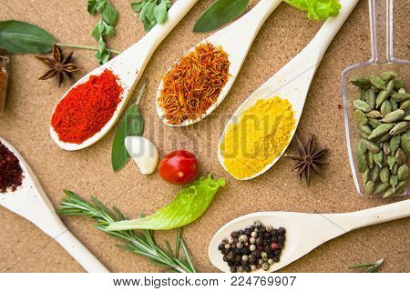 Cooking hot spicy meal. A set of four different spices in wooden spoons, dry beans, fresh cherry tomatoe, garlic, rosemary and salvia, corkwood background, top view