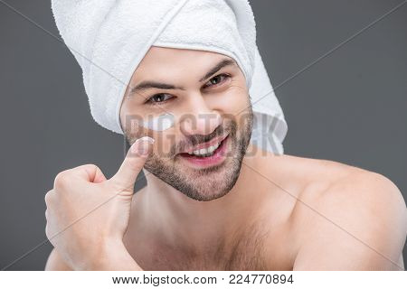 cheerful beardman in towel applying face cream, isolated on grey, skin care concept