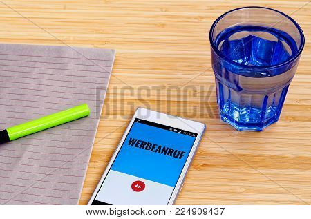 Mobile phone at which a advertising call and in German language stands "Werbeanruf" (in English: advertising call) with block and green pen and tumbler