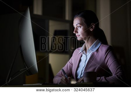 Serious Office Worker Looking At Computer Monitor And Making Notes At The Table She Working Till Lat