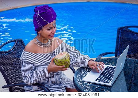 Afro American Girl Wearing An Ethnic Turban Near Swimming Pool . Indian Woman Drinking Coconut Water