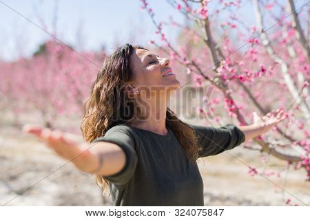 Beautiful middle age woman in the middle of pink peach flowers and trees smiling cheerful with open arms enjoying sunbathe on sunny day