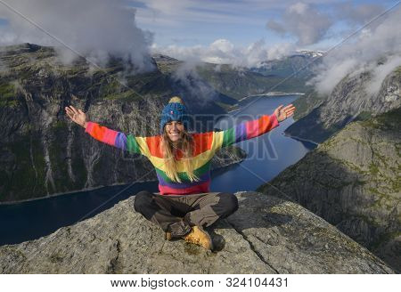 Girl With Outstreched Hands Sits On Rock Edge Against Blue Lake And Mountains Background.