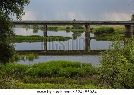 Bridge Over A Small River. Bridge Against A Cloudy Gray Sky With Rain. Rural Landscape.