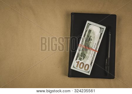 A Black Notebook , A Pen And Dollar Cash Banknotes On Wooden Background - Concept Of Financial Manag
