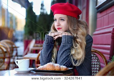 Girl In A Gray Coat And A Red Beret With Long Curls Sits Thoughtfully Near A Cafe On A Background Of