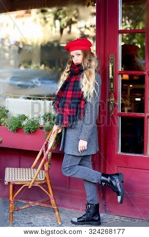 Girl In A Gray Coat And A Red Beret With Long Curls Stands Thoughtfully Near A Cafe On A Background 