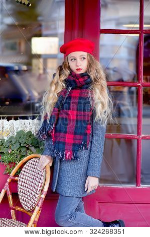 Girl In A Gray Coat And A Red Beret With Long Curls Stands Thoughtfully Near A Cafe On A Background 
