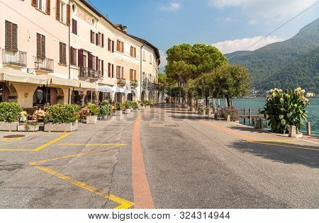 View Of Picturesque Village Morcote With Outdoor Bars And Gift Shops On The Shore Of Lake Lugano In