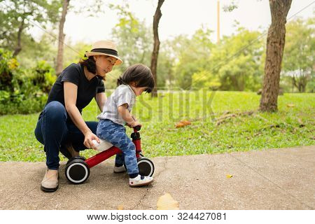 Young Asian Mother Teaching Her Adorable Toddler Girl To Ride The Bike For The First Time While Spen