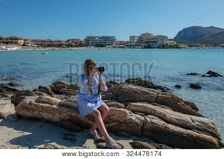 Woman In A Dress Sitting On The Rocks On The Beach And Taking Pictures With A Slr Camera. Sea And Mo