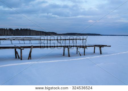 Old Wooden Pier In The Snow. Frozen Lake And Pine Forest. Countryside