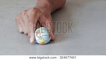 Small Globe Held By Fingers With Grey Concrete Background Seen From The Front. World In Our Hands Wi