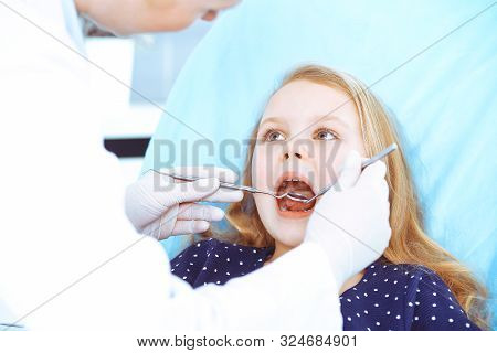 Little Baby Girl Sitting At Dental Chair With Open Mouth During Oral Check Up While Doctor. Visiting