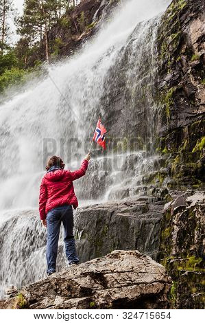 Tourist Woman With Norwegian Flag At Svandalsfossen In Norway, Powerful Waterfall In Norwegian Mount