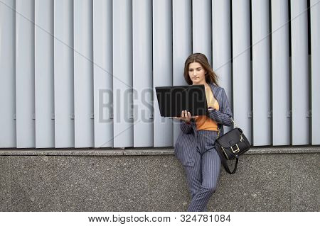 Young Businesswoman With Laptop Stay In The City Against The Background Of The Wall, A Girl In A Str