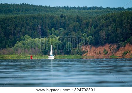 Sailing boat motoring on Kama river at calm windless day with steep coast and forest on the background. Russia