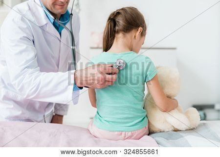 Young pediatrician in whitecoat examining back of little girl with stethoscope in medical office