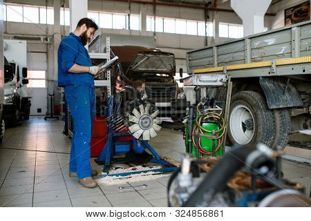 Young serious worker of automobile repair service reading instructions of transport maintenace while standing in workshop