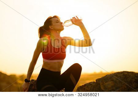 Young Woman Resting and Drinking Water after Trail Running Outdoor. Workout Exercise at Hot Beautiful Summer Sunset. Sport and Healthy Active Lifesyle Concept.
