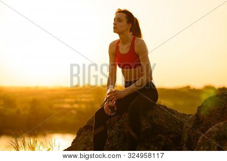 Young Woman Resting and Drinking Water after Trail Running Outdoor. Workout Exercise at Hot Beautiful Summer Sunset. Sport and Healthy Active Lifesyle Concept.