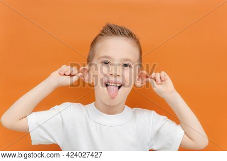 A Kid Makes A Face On An Orange Background. Caucasian Cheerful Boy Shows His Tongue And Pulls Back H
