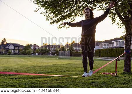 Adventurous White Caucasian Adult Woman Walking On A Slackline Between Trees In A Neighborhood Park.