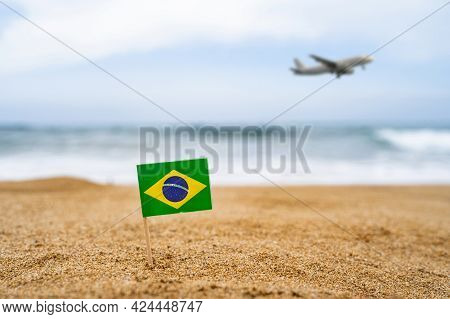 Flag Of Brazil In The Form Of A Toothpick In The Sand Of Beach Opposite Sea Wave With Landing Airpla