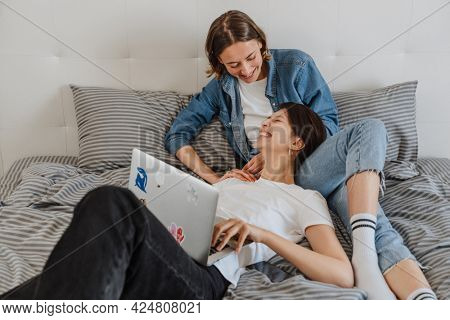 A laughing lesbian couple with laptop looking to each other while lying on the bed in the light room