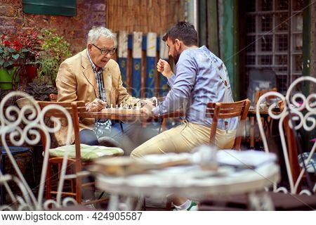 young beardy guy playing chess with elderly man, sitting in outdoor cafe. selective focus image