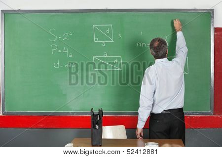 Rear view of mature male teacher writing on greenboard in classroom