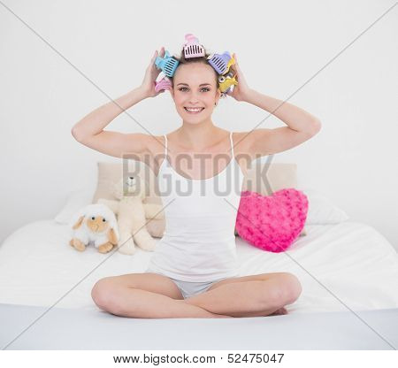 Pretty natural brown haired woman fixing her hair curlers in bright bedroom
