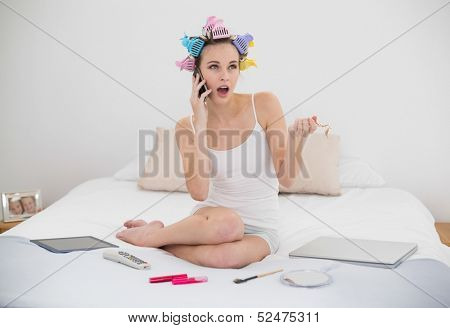 Shocked natural brown haired woman in hair curlers calling with her mobile phone in bright bedroom
