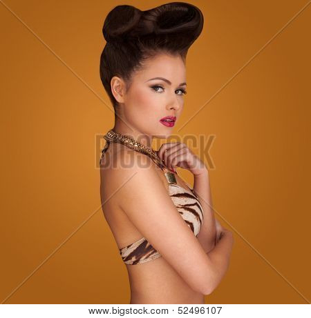 Beautiful pin up girl with a glamorous swept up hairdo wearing gold jewellery posing in an animal print bikinin against a brown studio background