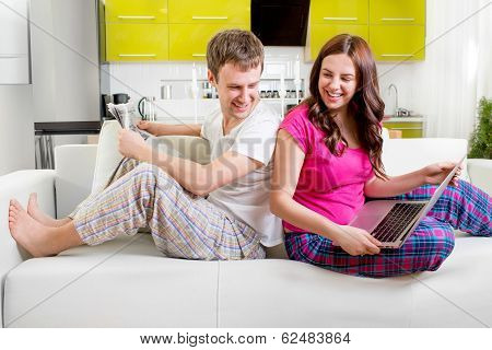 Young Married Couple In Pajamas Sitting In The Sofa Reading Newspaper And Laughing At Home In The Mo