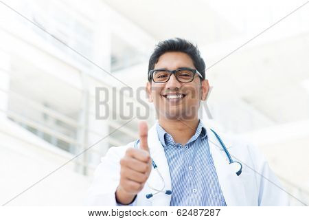Portrait of a smiling Asian Indian male medical doctor in uniform thumb up, hospital building at background.