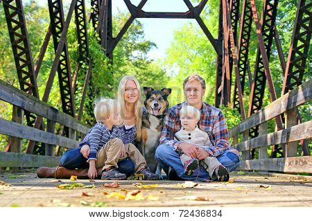 Family Of Four People And Dog Sitting On Bridge In Autumn