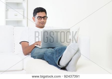 Young Indian guy doing online business with laptop computer at home. Asian man using internet indoor, relaxed and sitting on sofa. Handsome male model.