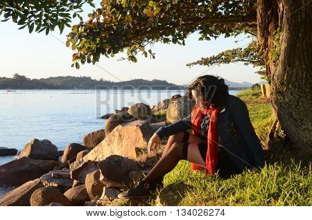 Young white woman, black and curly hair, sitting in grass at beach landscape