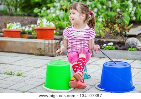 Little girl having fun in the summer garden pretending that she plays the drums using a plastic bucket